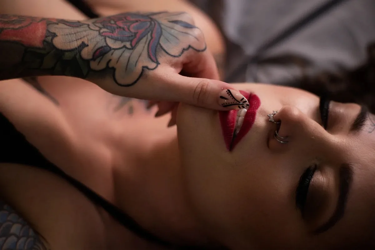 Boudoir portrait of a woman lying down with a tattooed arm raised to her face, showing patterned nail art on a manicured fingernail.